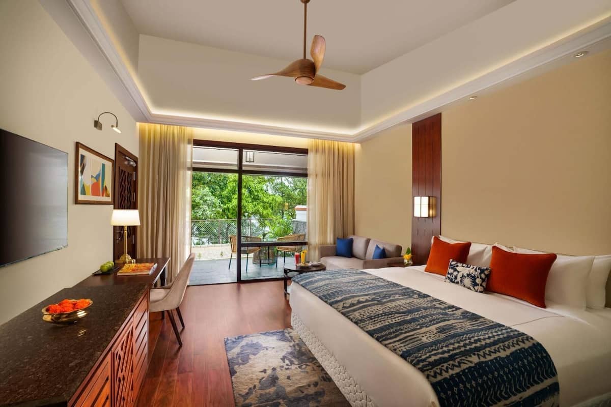 A hotel room with a bed, a desk, a chair, a TV, a balcony with a view of greenery, and a ceiling fan.