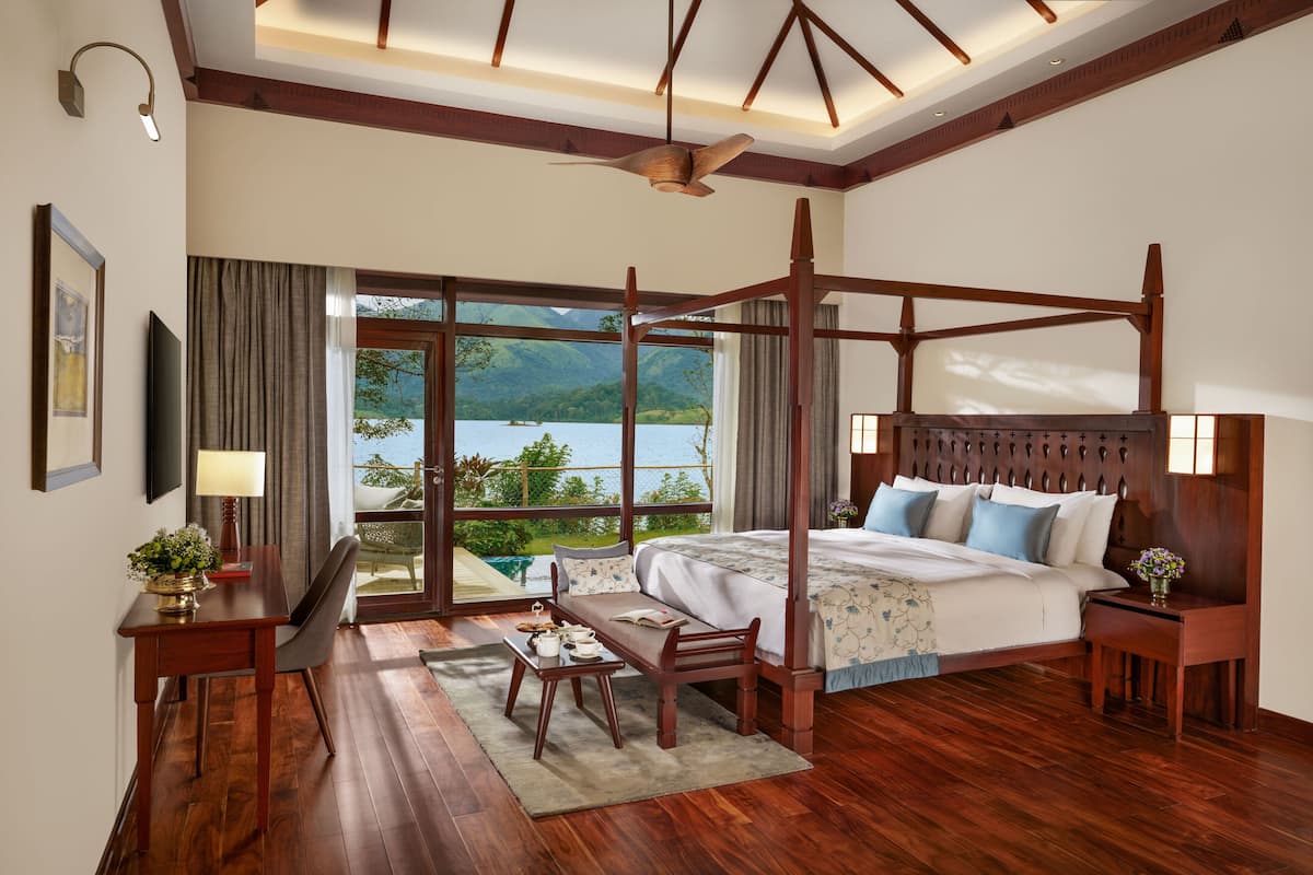 A spacious bedroom with a four-poster bed, a wooden desk, and a view of a lake.