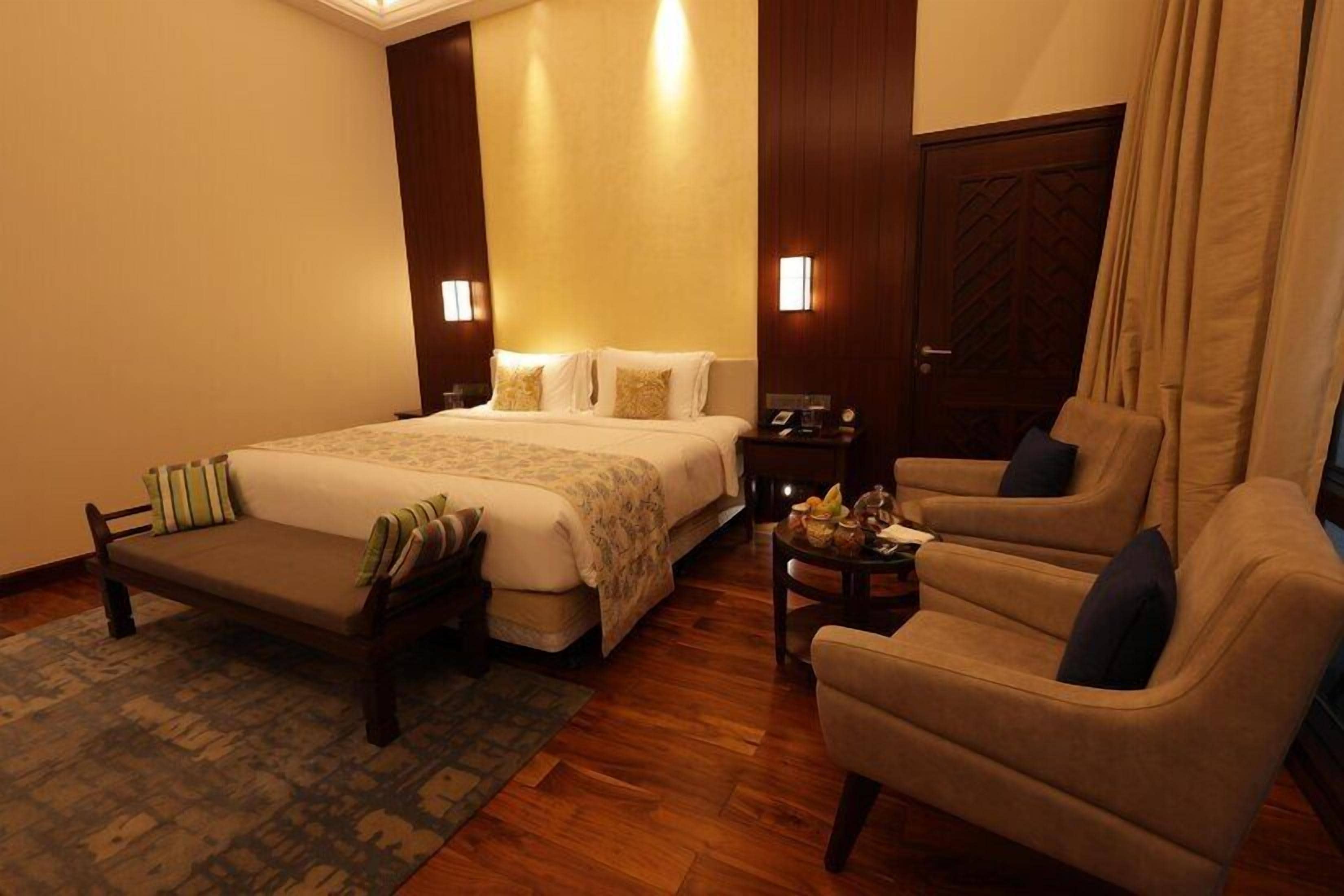 A hotel room with a large bed, a sofa, a chair, a small table with a lamp, and a wooden door.