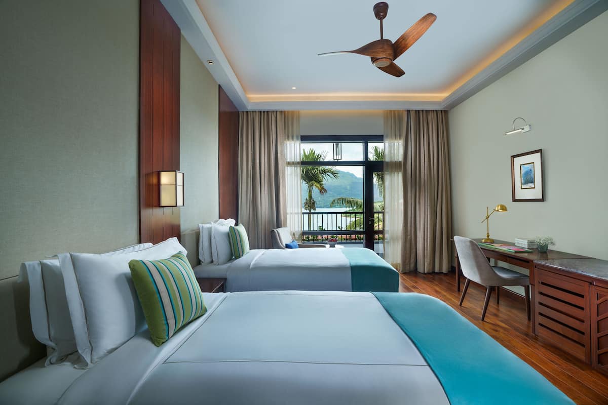 A hotel room with two beds, a desk, a chair, a ceiling fan, and a view of the outdoors.