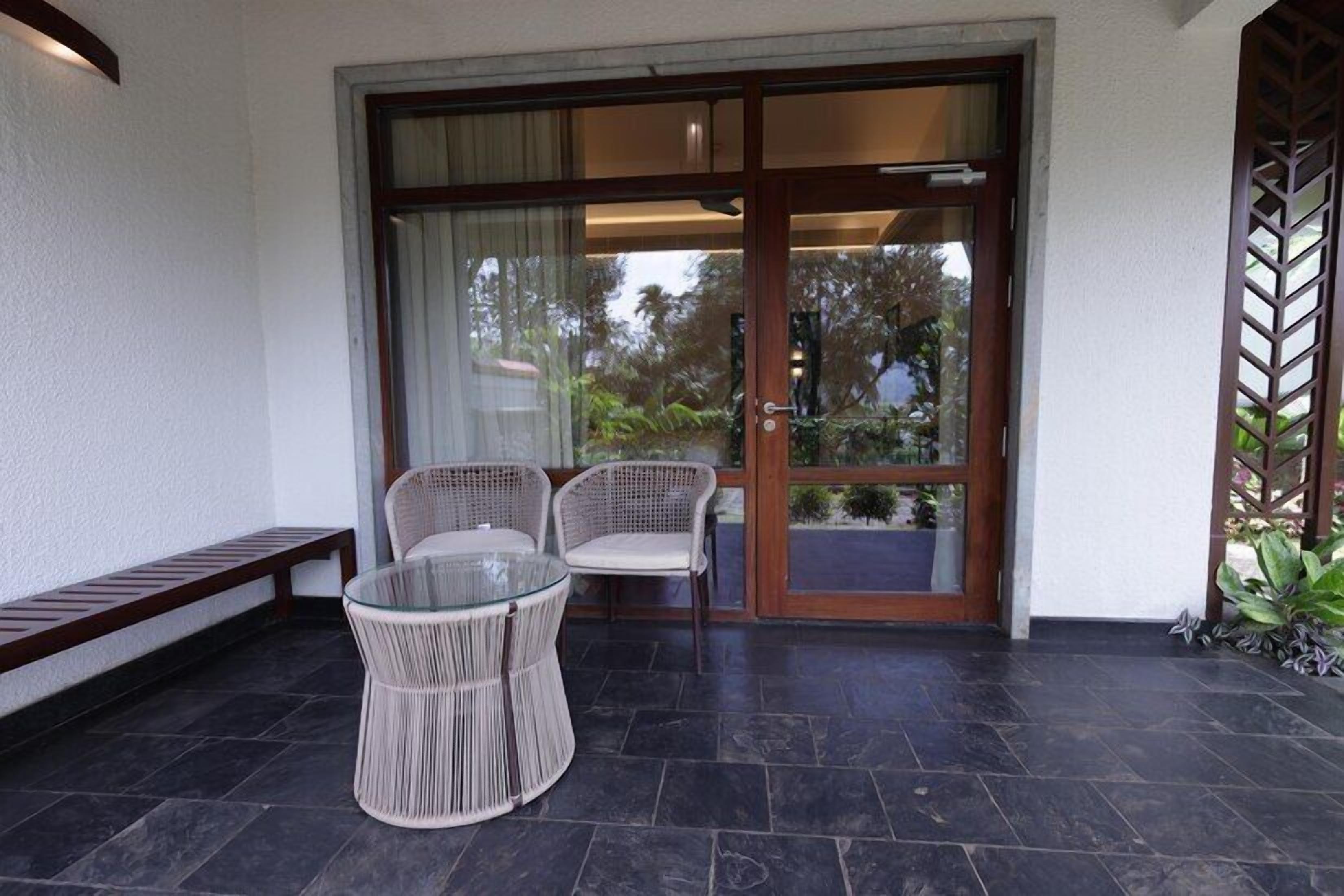 A covered outdoor seating area with a glass table and wicker chairs, a sliding glass door leading to a garden, and a bench along the wall.
