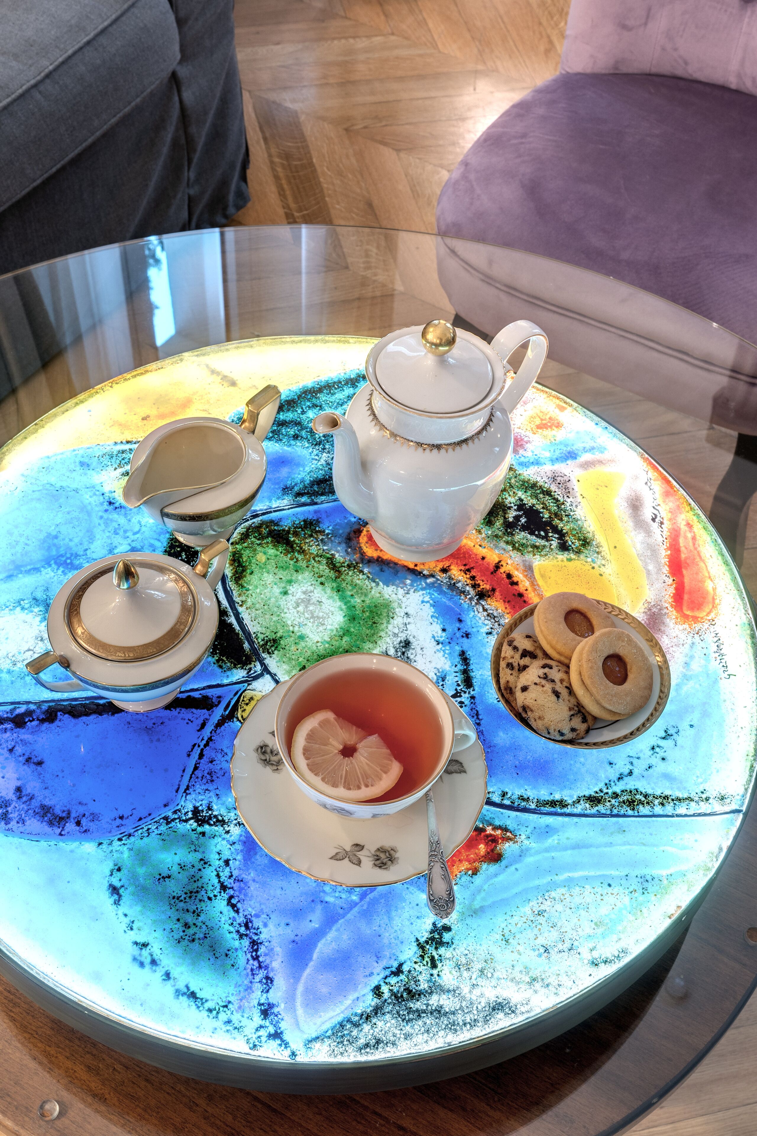 A round glass table with a colorful marbled surface, a white teapot, two cups, a teapot lid, a teapot strainer, and two cookies.