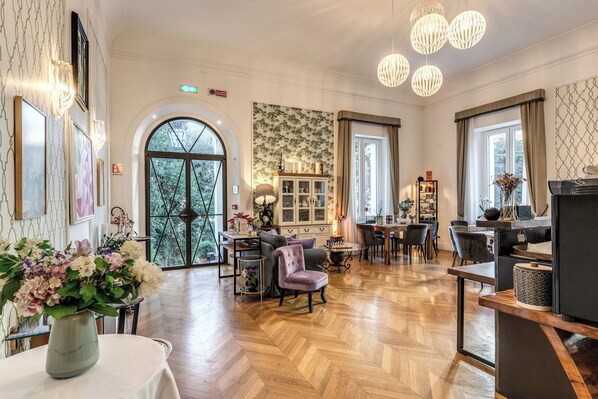 Un hall d’hôtel spacieux avec un parquet en bois disposé en motif chevron, un coin salon avec un canapé et des fauteuils, un espace repas avec une table et des chaises, et un comptoir d’accueil.