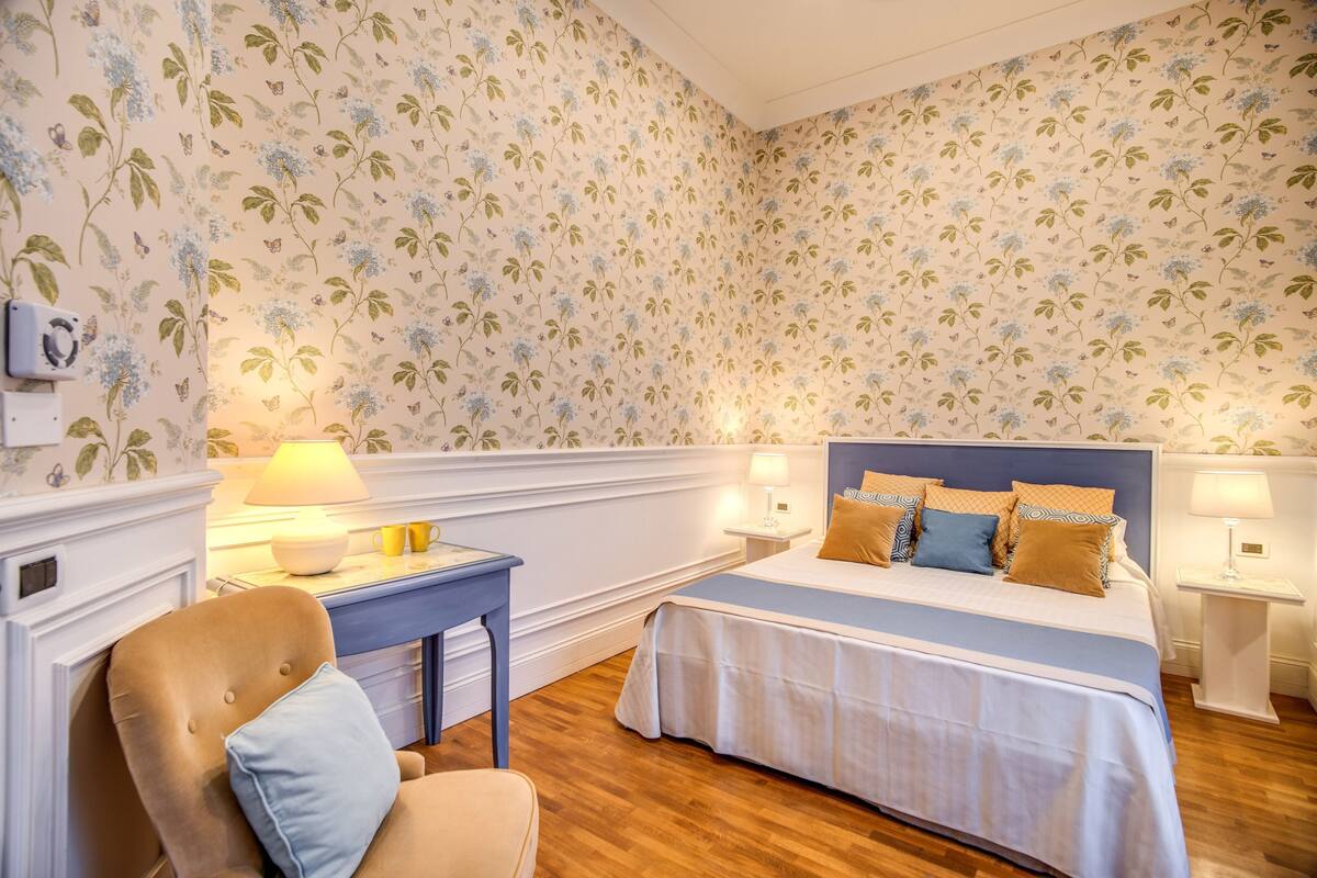 A bedroom with a bed, bedside tables, a chair, and floral wallpaper.