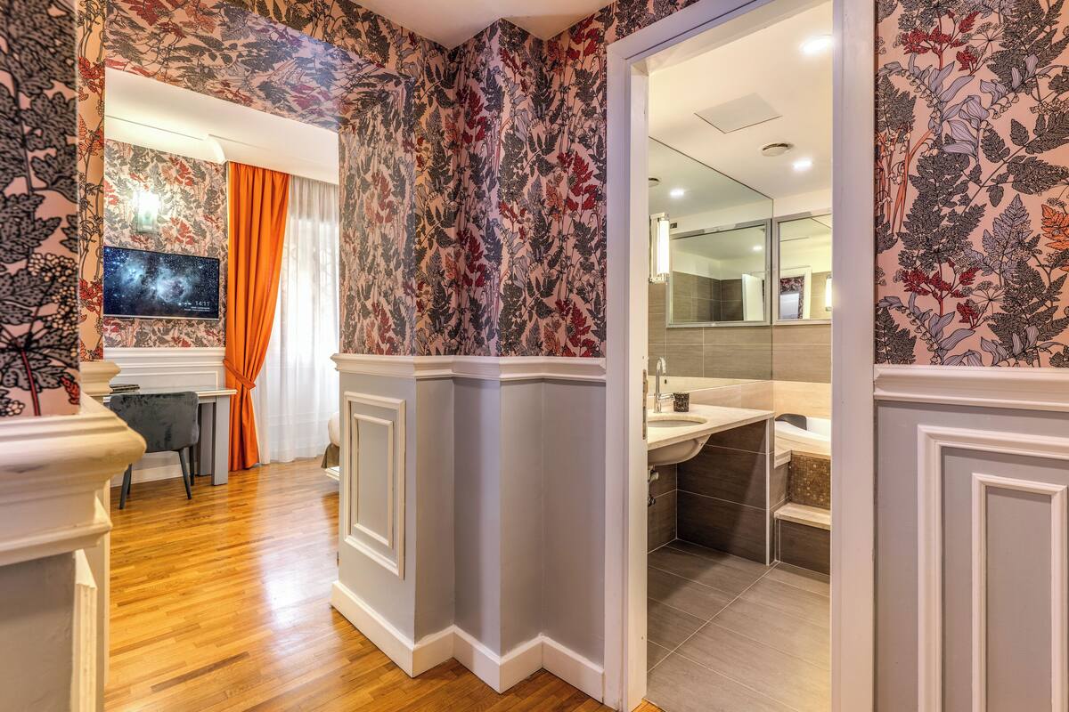 A room with a wooden floor, a television, a desk, and a bathroom with a sink and mirror.