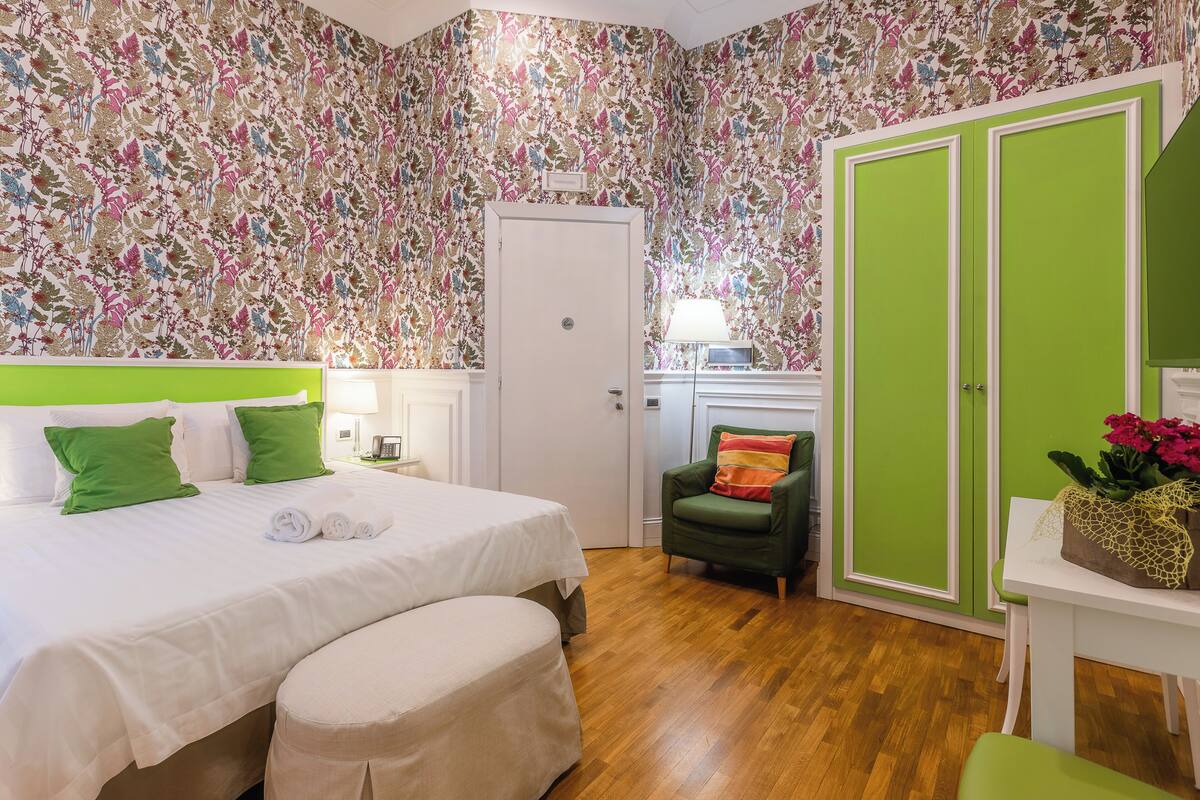 A bedroom with a floral wallpaper, a green wardrobe, a green armchair, and a white bedside table with a flower vase.