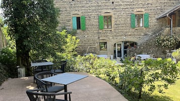 A stone building with green shutters, outdoor seating with tables and chairs, and a garden area with plants and trees.