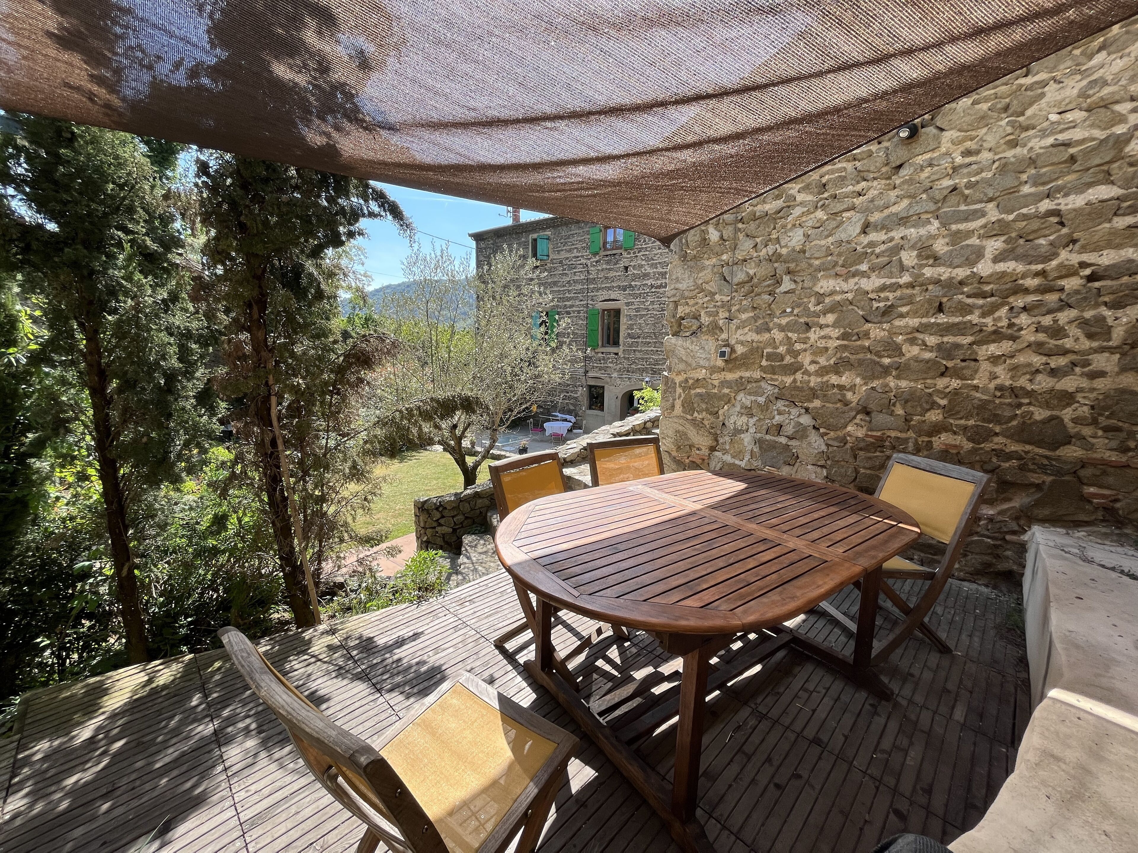 Une terrasse en bois avec une table et des chaises, un mur en pierre et une vue sur un bâtiment aux volets verts.