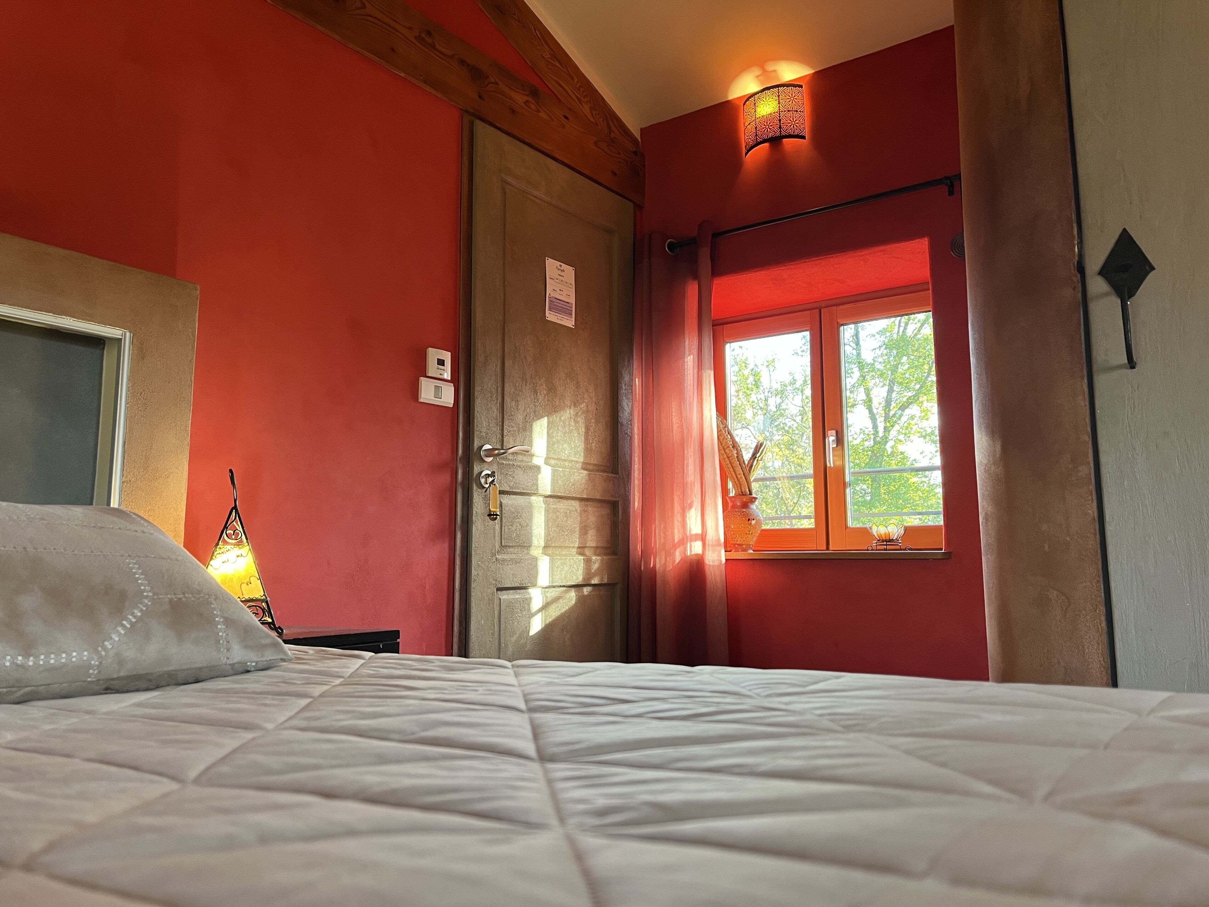Ein Schlafzimmer mit Bett, Fenster mit Blick auf Bäume, einer Tür und einer Wandleuchte.
