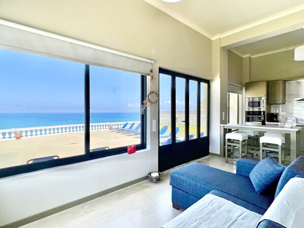 A room with a large window overlooking a beach, a kitchenette, and a seating area with a sofa and chairs.