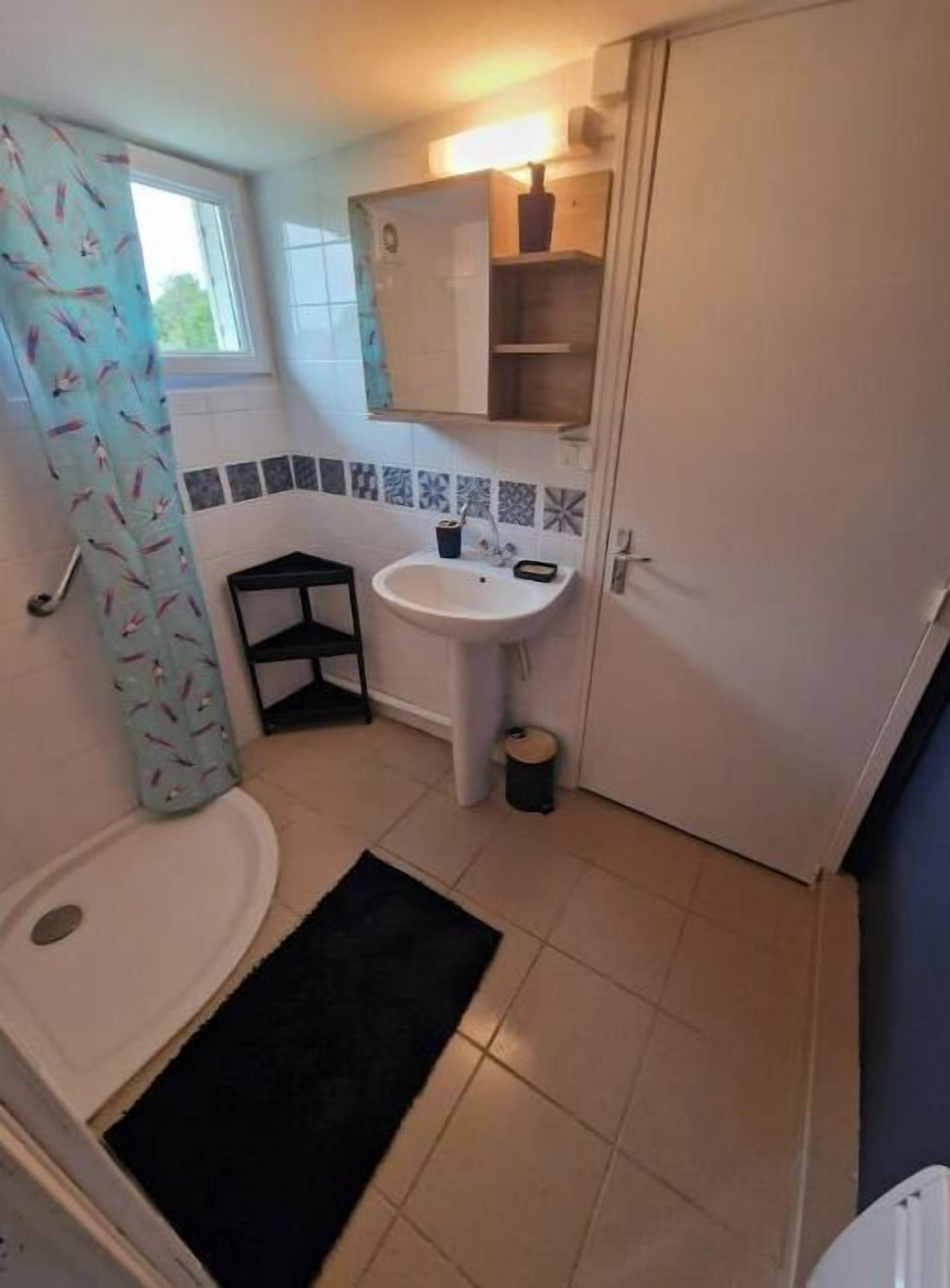 Un baño pequeño con un lavamanos blanco, un espejo y una ducha. Hay una ventana con cortina, y el piso está cubierto de azulejos.