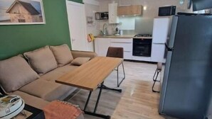 A compact living space with a sofa, a small dining table, a kitchen area with a refrigerator, microwave, and cabinets. - Small cottage (Tourville-sur-Sienne)