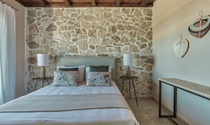 A bedroom with a stone wall, a bed with patterned pillows, two bedside lamps, and a small round table.