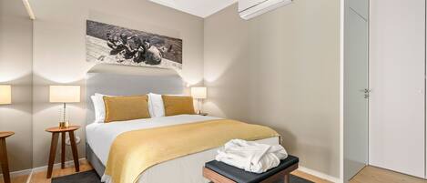 Un cuarto de hotel ordenado, con una cama grande, una mesita de noche con una lámpara, un banco con una toalla y un cuadro en la pared.
