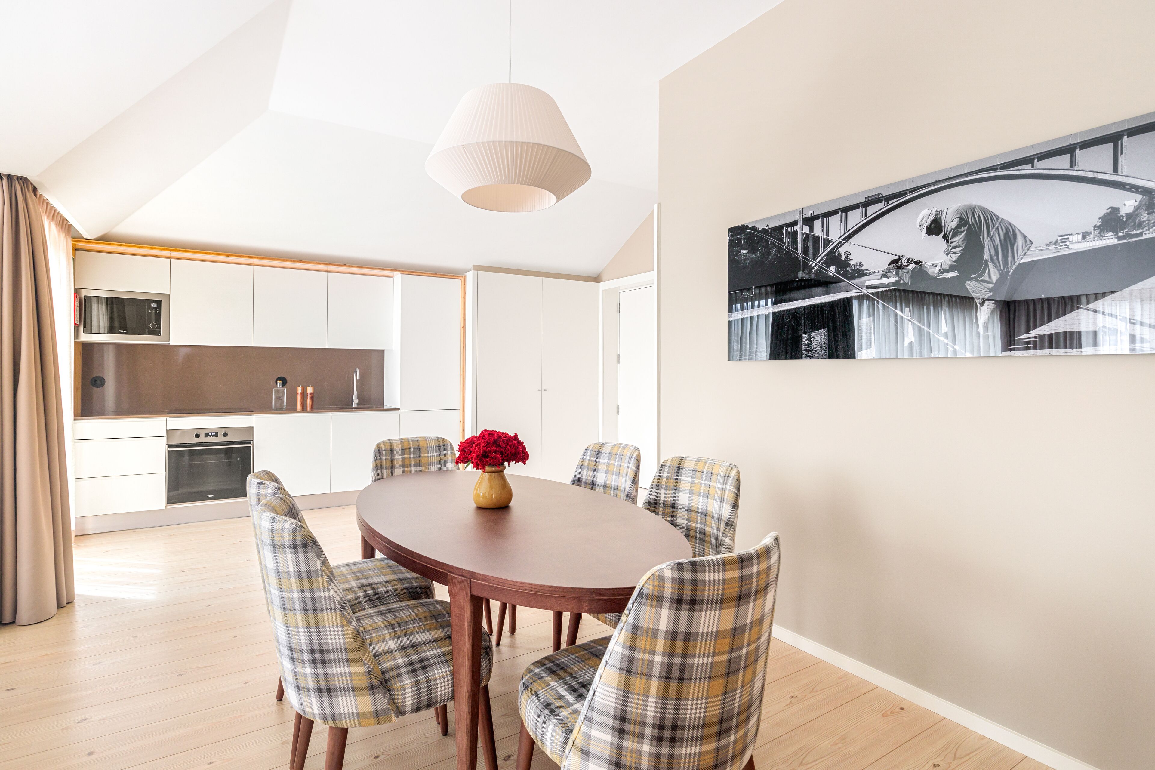A modern kitchen with white cabinets, a microwave, and a stove. There is a dining table with plaid chairs and a vase of red flowers.