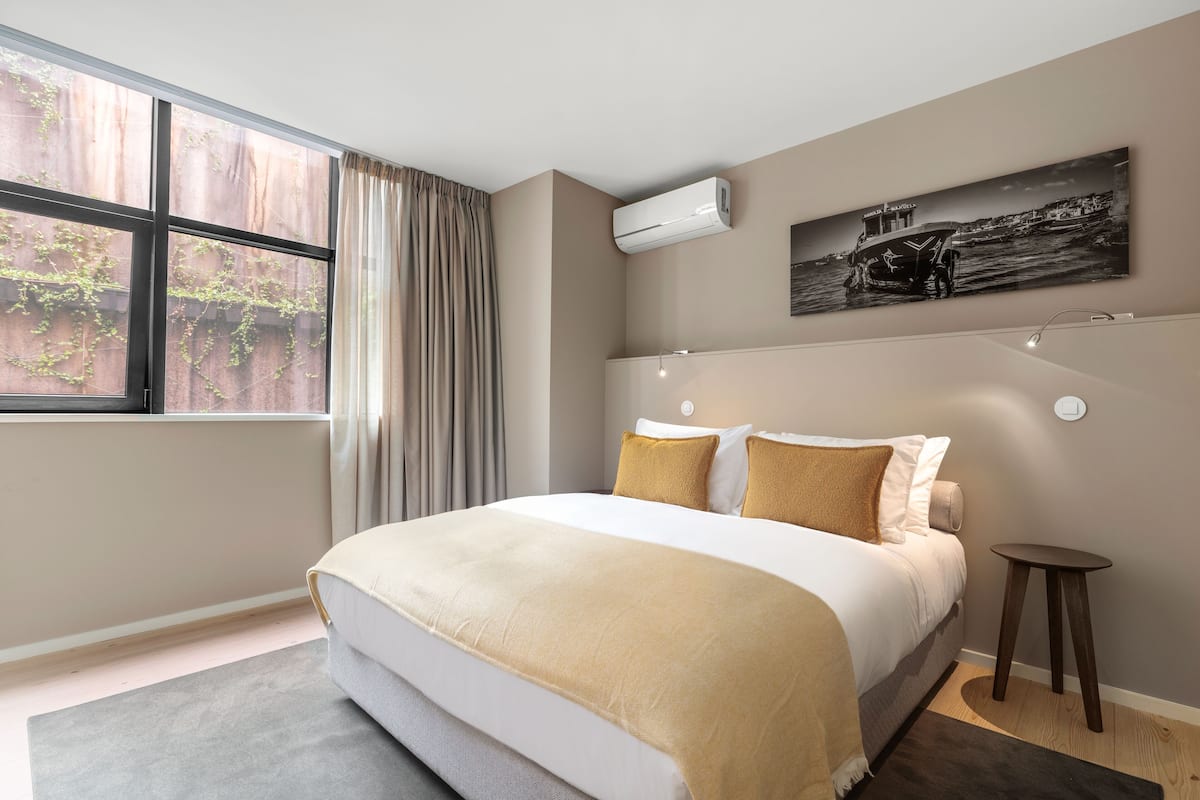 A modern bedroom with a large bed, a window with curtains, an air conditioning unit, a small table, and a wall-mounted artwork.