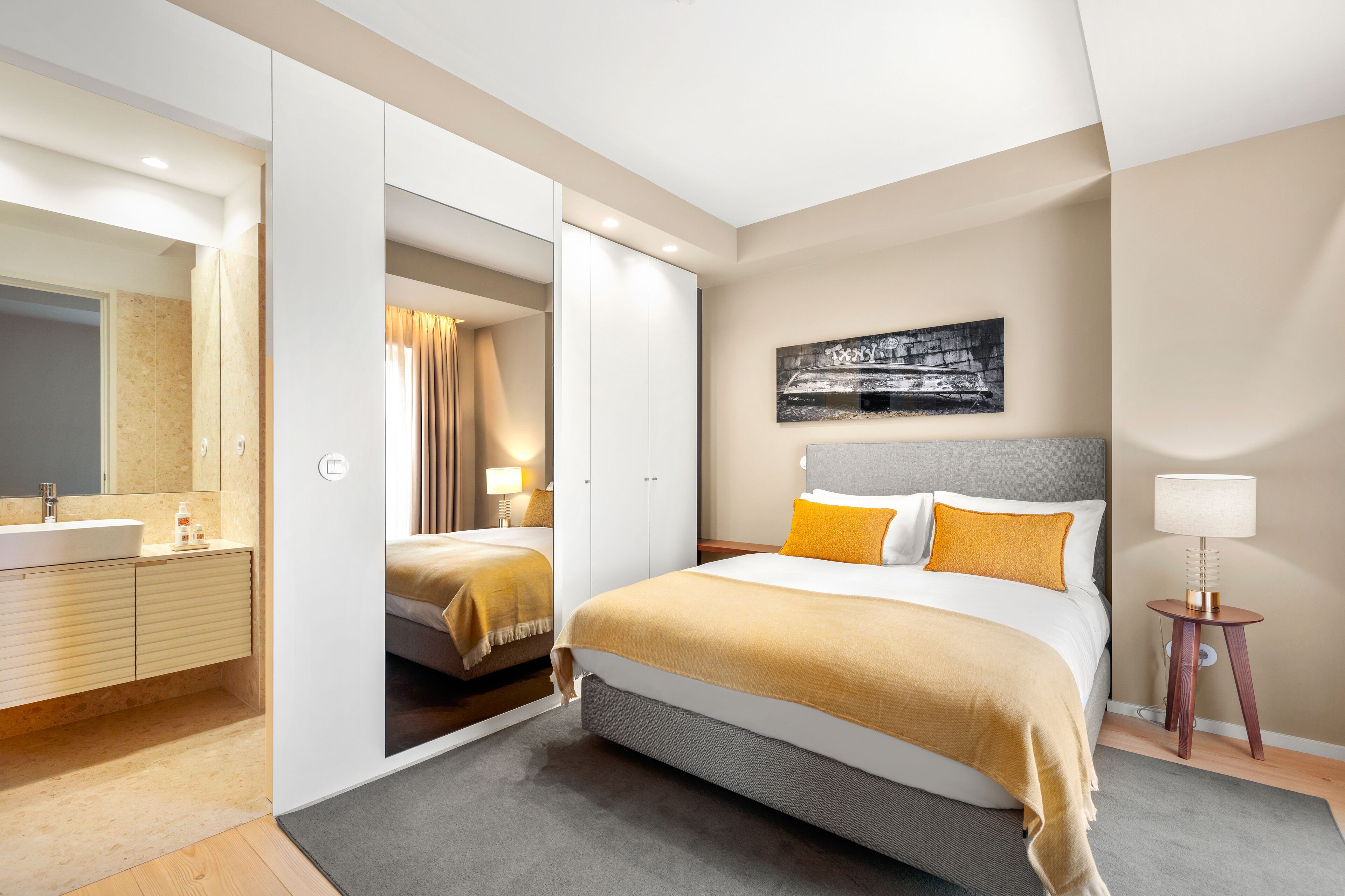 A modern bedroom with a large bed, a walk-in closet, a bathroom with a sink, and a wall-mounted artwork.