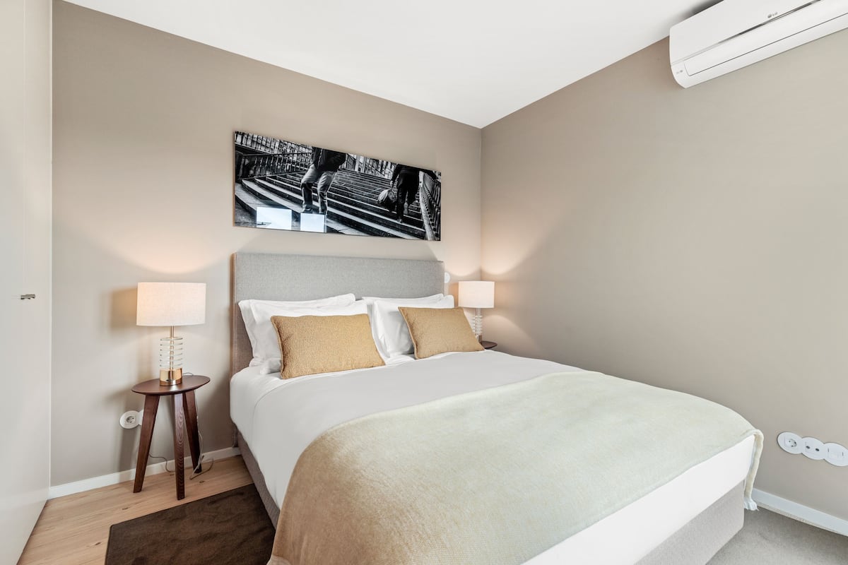 A neatly arranged bedroom with a bed, two bedside lamps, a wall-mounted air conditioner, and a framed picture above the bed.