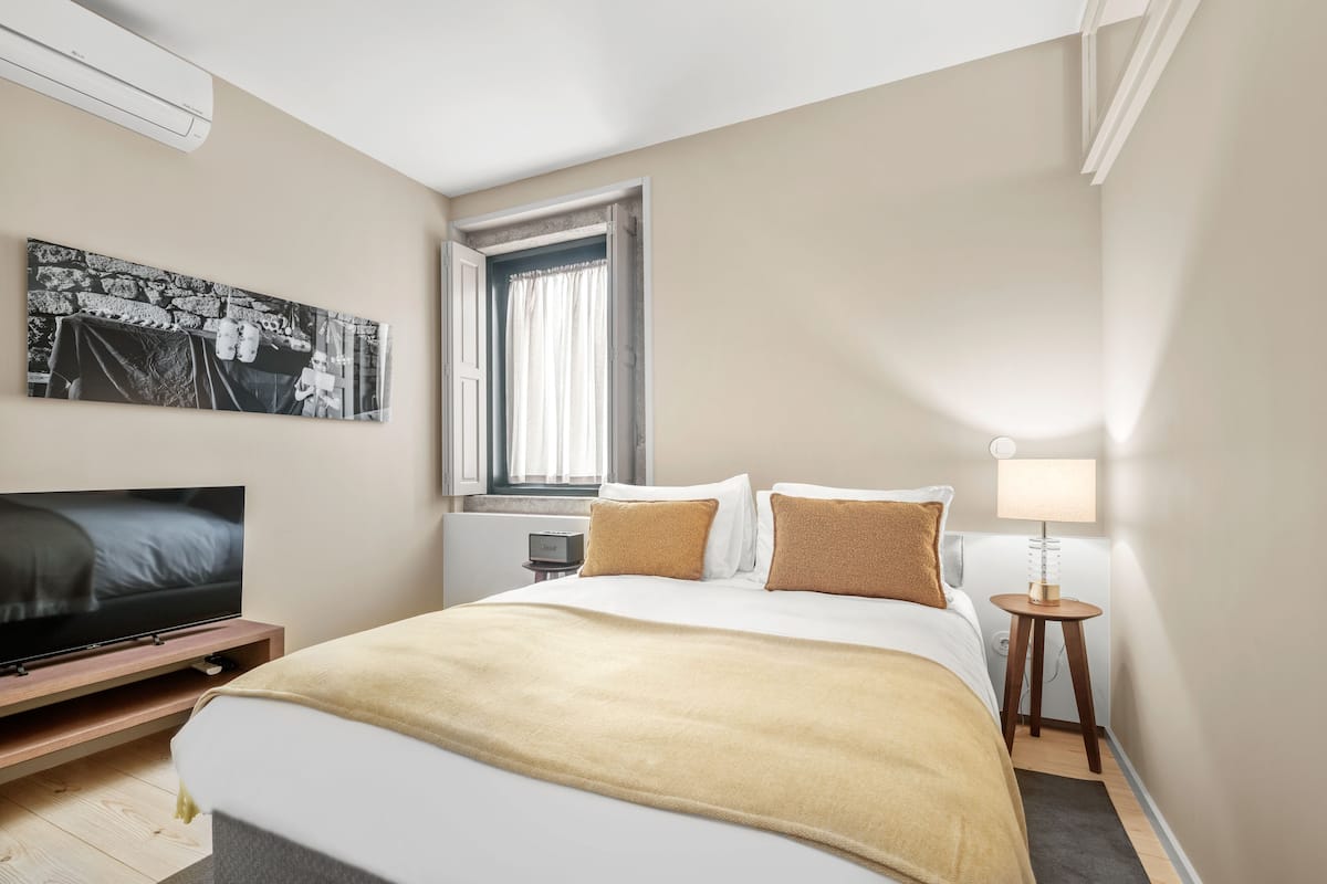 A modern hotel room with a bed, a flat-screen TV, a window with curtains, a bedside table with a lamp, and a wall-mounted artwork.
