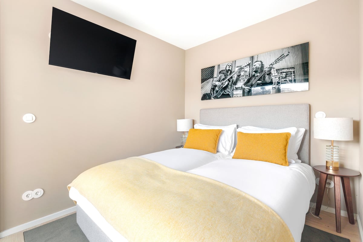 A hotel room with a large bed, two bedside lamps, a flat-screen TV mounted on the wall, and a framed black-and-white photograph above the bed.