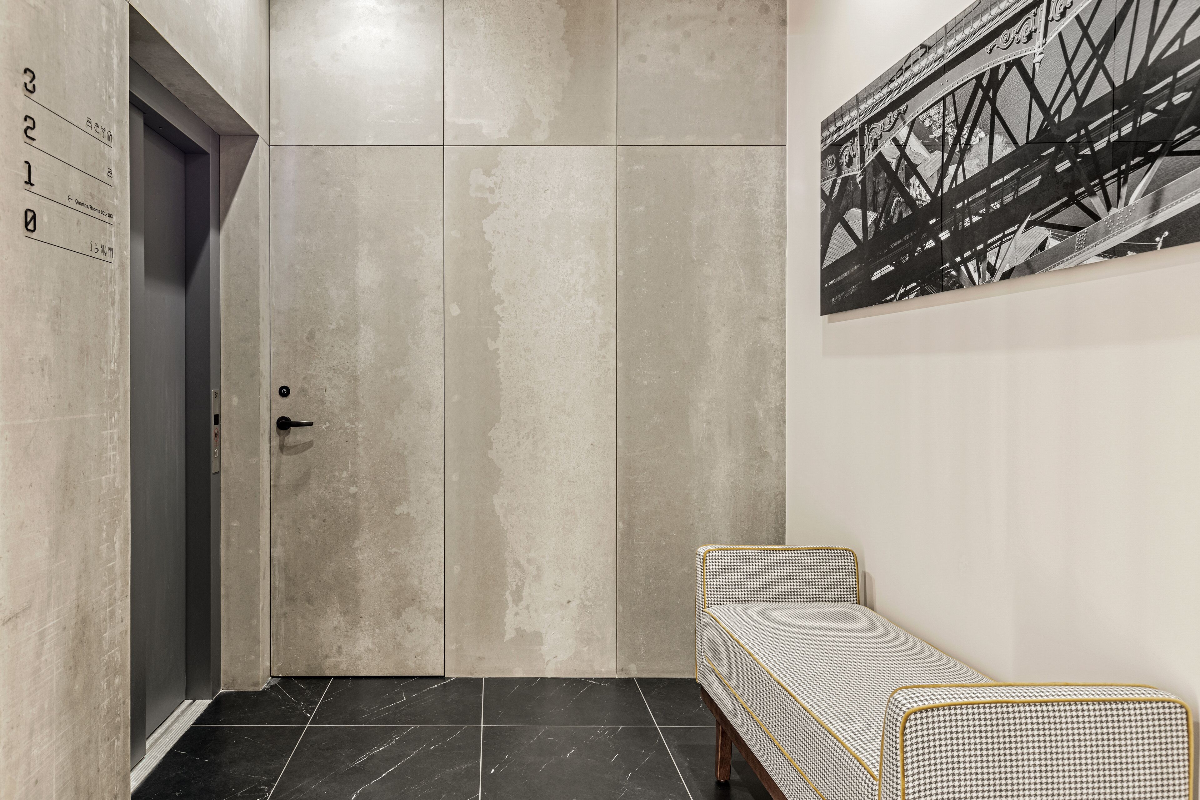 Un ascenseur moderne avec une porte grise, un panneau de numéros fixé au mur et un banc à motifs noir et blanc.