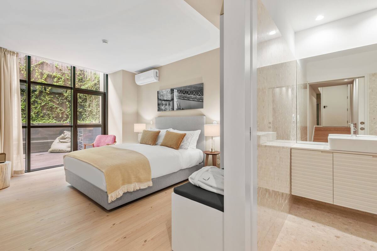 A modern bedroom with a large bed, a bedside table, a nightstand, a window with a view of greenery, and a bathroom with a mirror and sink.