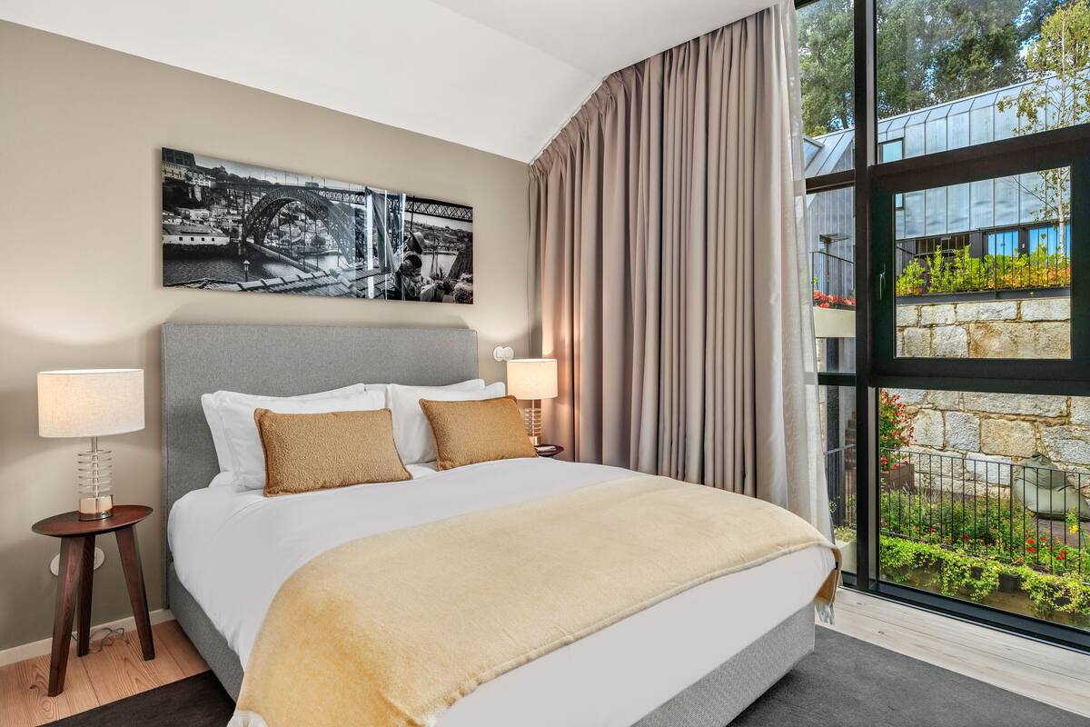 A modern hotel room with a large bed, two bedside tables, a nightstand, a large window with curtains, and a cityscape view.