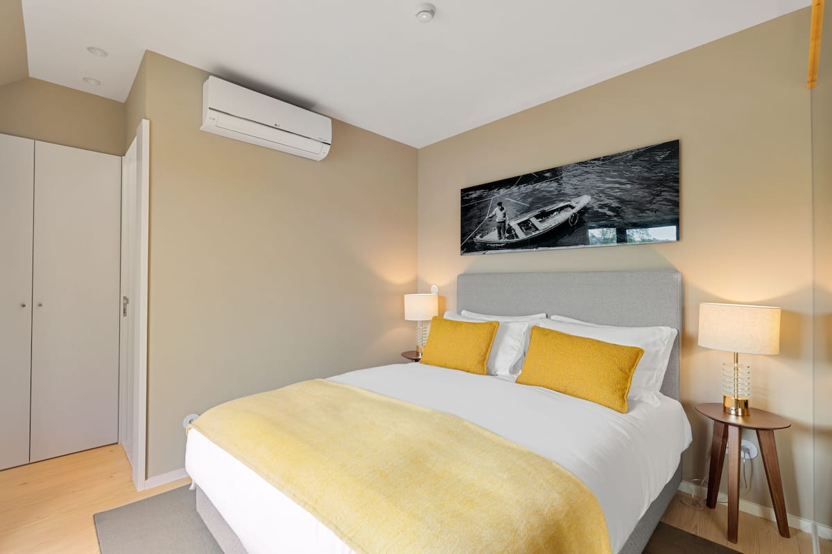 A hotel room with a bed, bedside lamps, a painting on the wall, and an air conditioning unit.