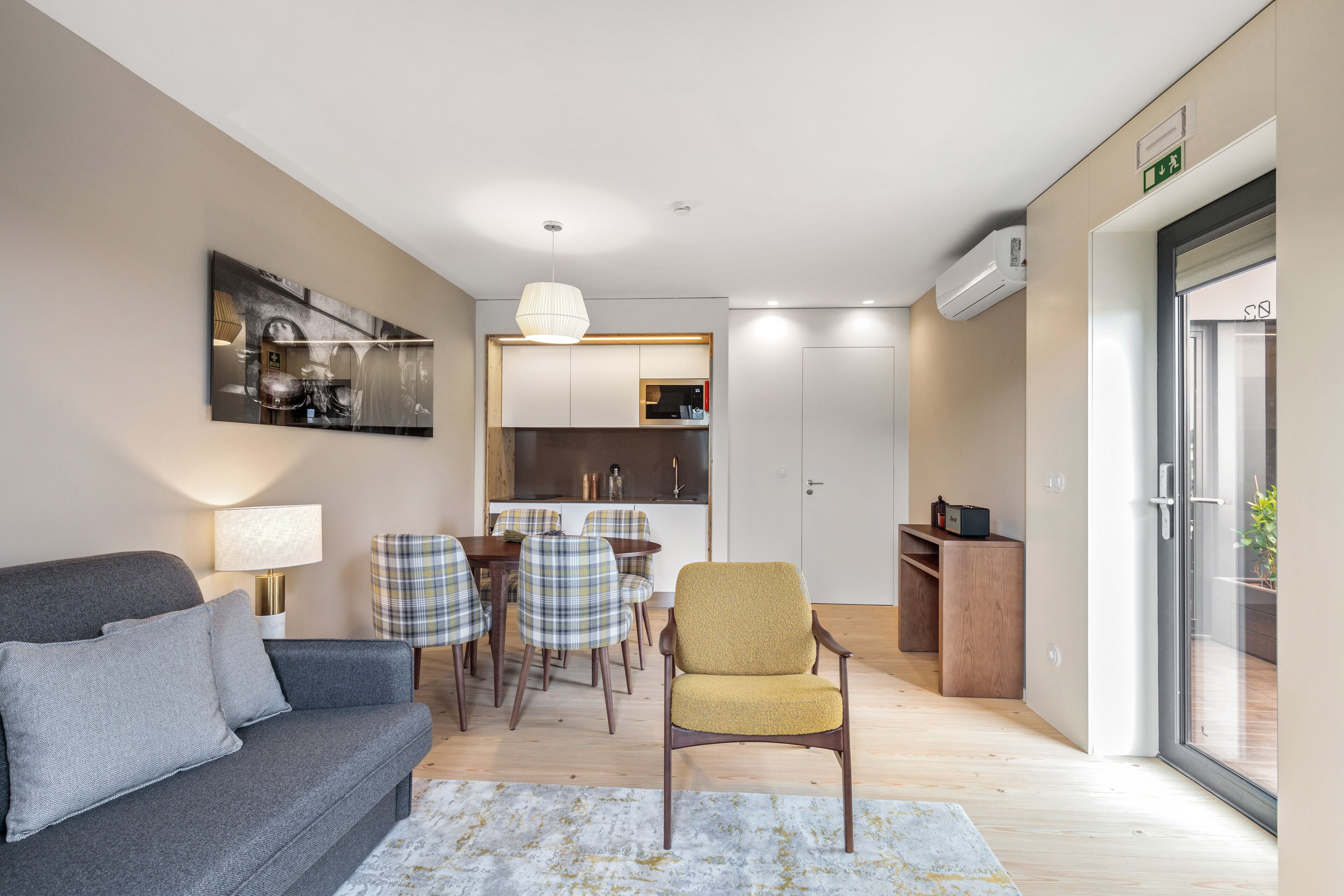 Ein modernes Wohnzimmer mit einer Couch, einem gelben Sessel, einem Esstisch mit karierten Stühlen und einer Küchenzeile mit einer Pendelleuchte.