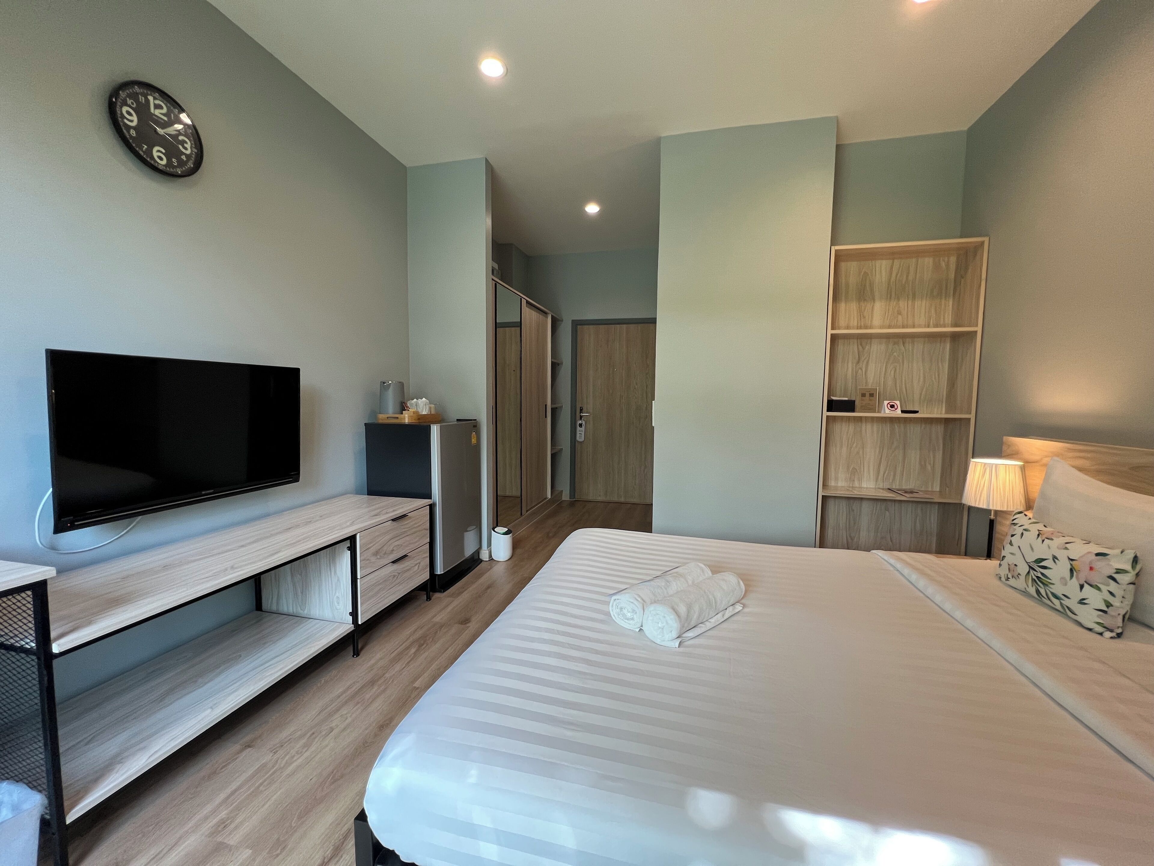 Une chambre d’hôtel moderne équipée d’une télévision à écran plat, d’un bureau en bois, d’un lit avec une couette blanche, d’une armoire en bois et d’une table de chevet avec une lampe.