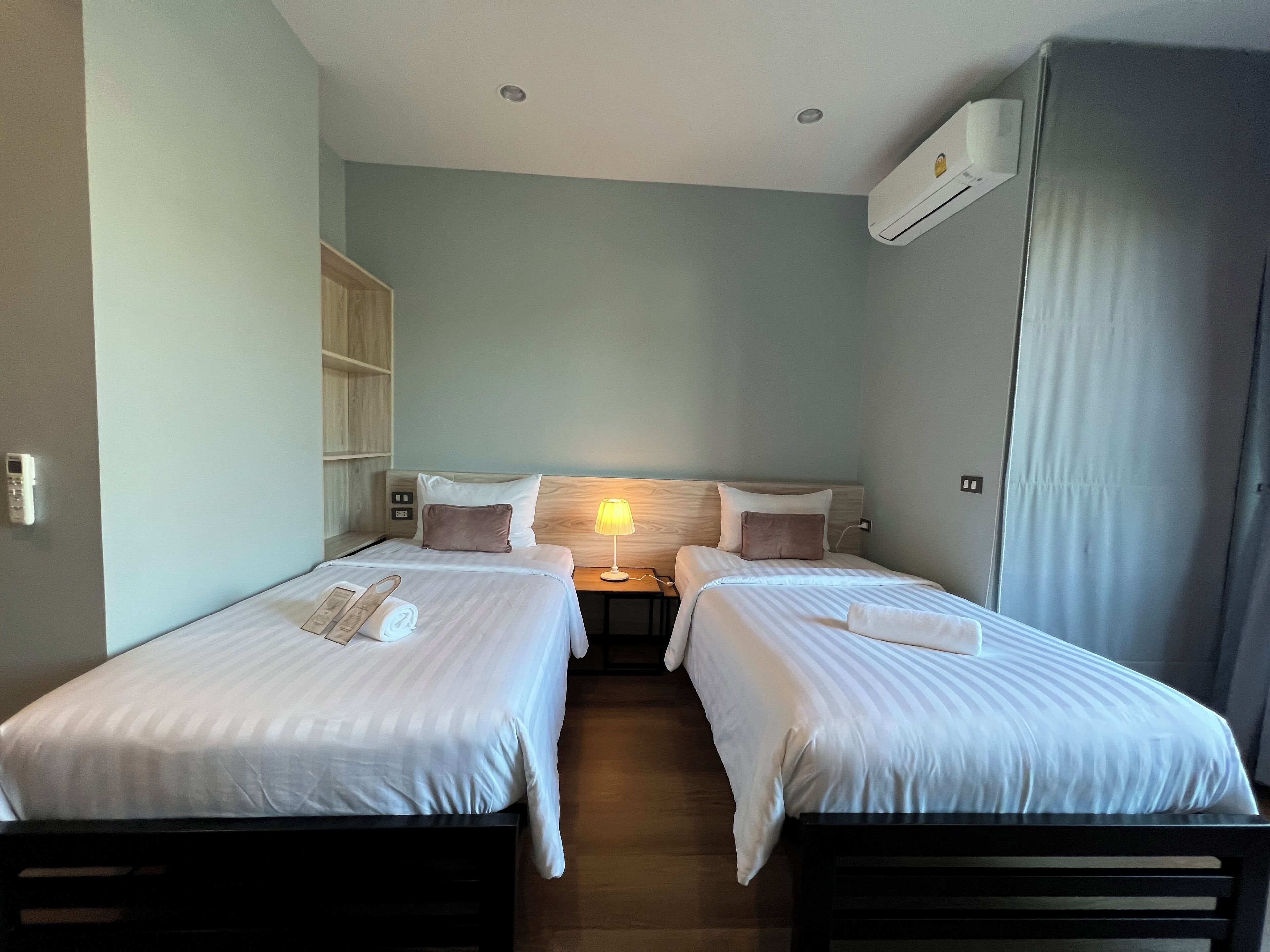 A hotel room with two single beds, a bedside table with a lamp, a wooden headboard, and an air conditioning unit.