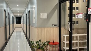 Un pasillo de hotel con un largo suelo de baldosas reflectante, una cesta de mimbre, una pequeña mesa roja y una puerta de cristal que da a una habitación con cama y escritorio.