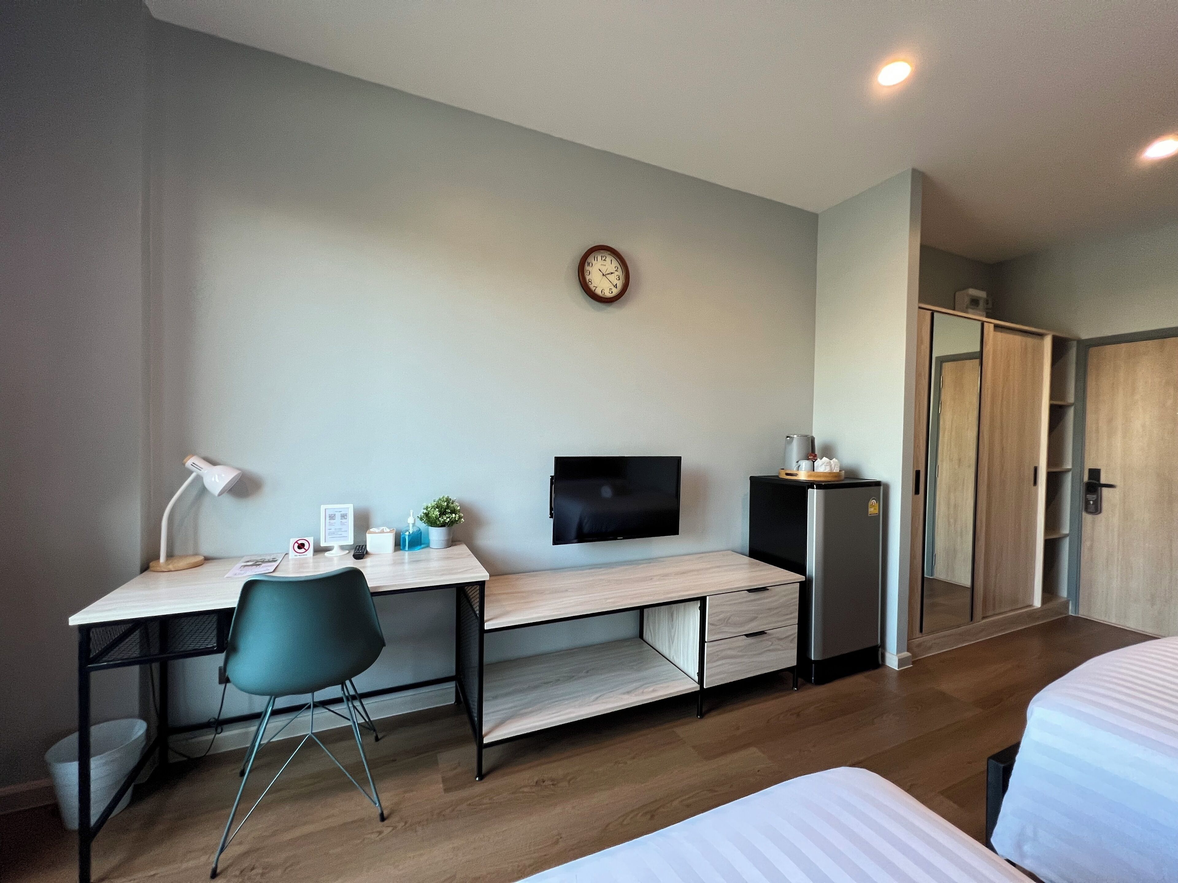 Een moderne hotelkamer met een houten bureau, een stoel, een bed, een televisie, een kleine koelkast en een klok aan de muur.