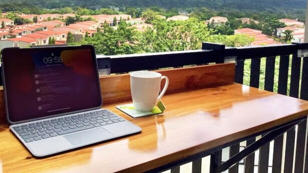 A laptop on a wooden table with a white mug, overlooking a residential area with red-roofed houses and green hills in the background. - Near everything! (Santa Tecla)