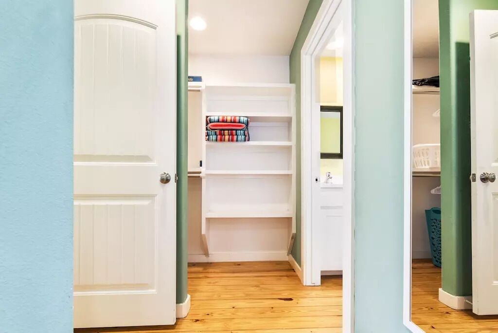 Un couloir au plancher en bois menant à une porte blanche, un placard avec des étagères et un panier à linge.