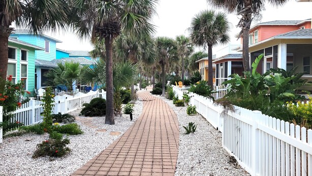 A pathway lined with white picket fences, palm trees, and a variety of plants. - Village Walk 3 BR/2.5 Bath--Shared Heated Pool and Close to the Beach! (Port Aransas)