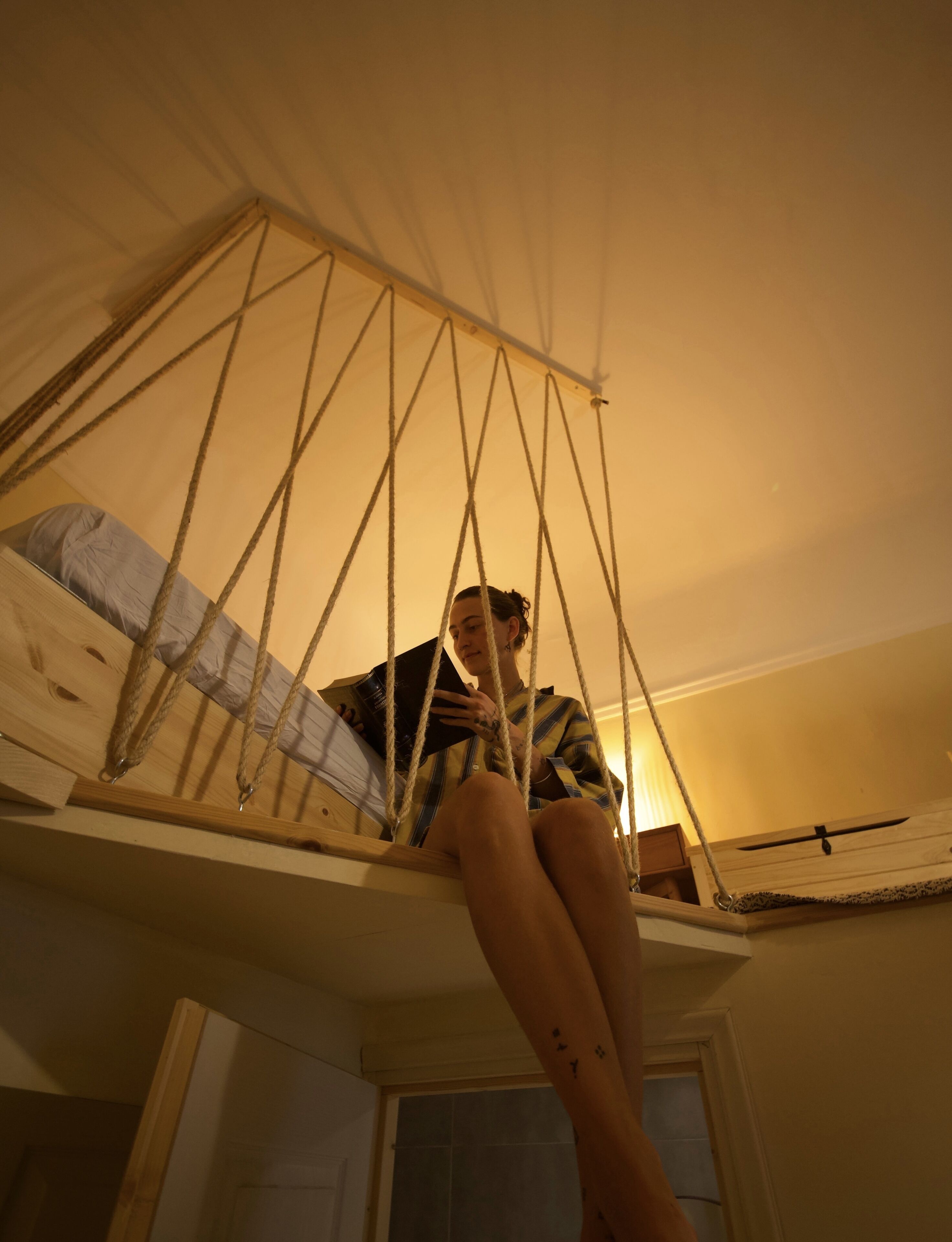 A person sitting on a suspended wooden platform with ropes, reading a book.