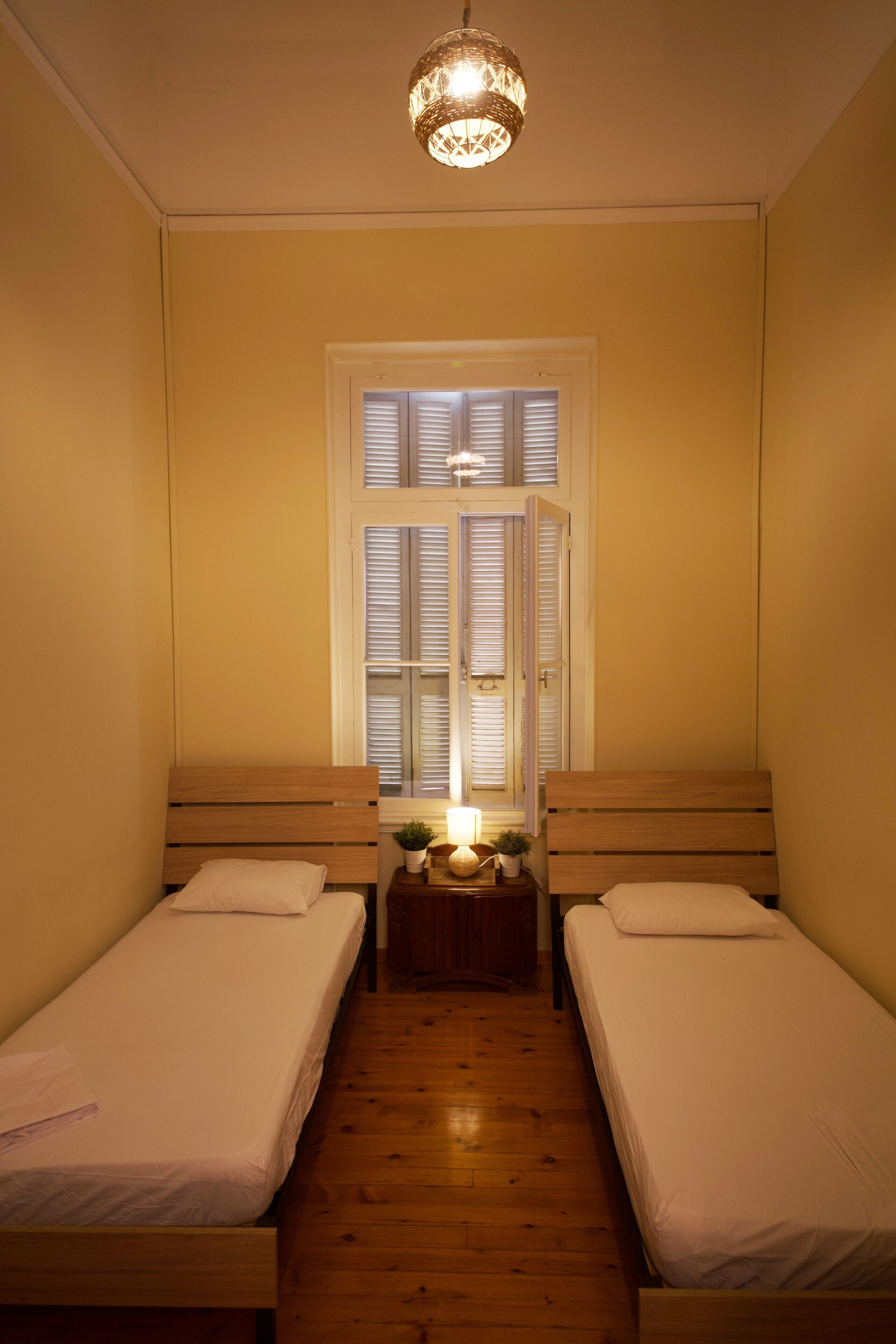 Una habitación pequeña y acogedora con dos camas individuales, piso de madera, una ventana con contraventanas y una lámpara colgante.