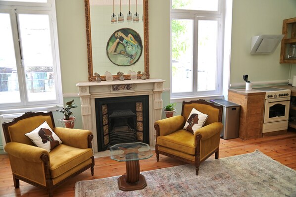 A living room with two yellow armchairs, a fireplace, a painting, and a kitchen area with appliances. - Baloo Hostel (Athens)