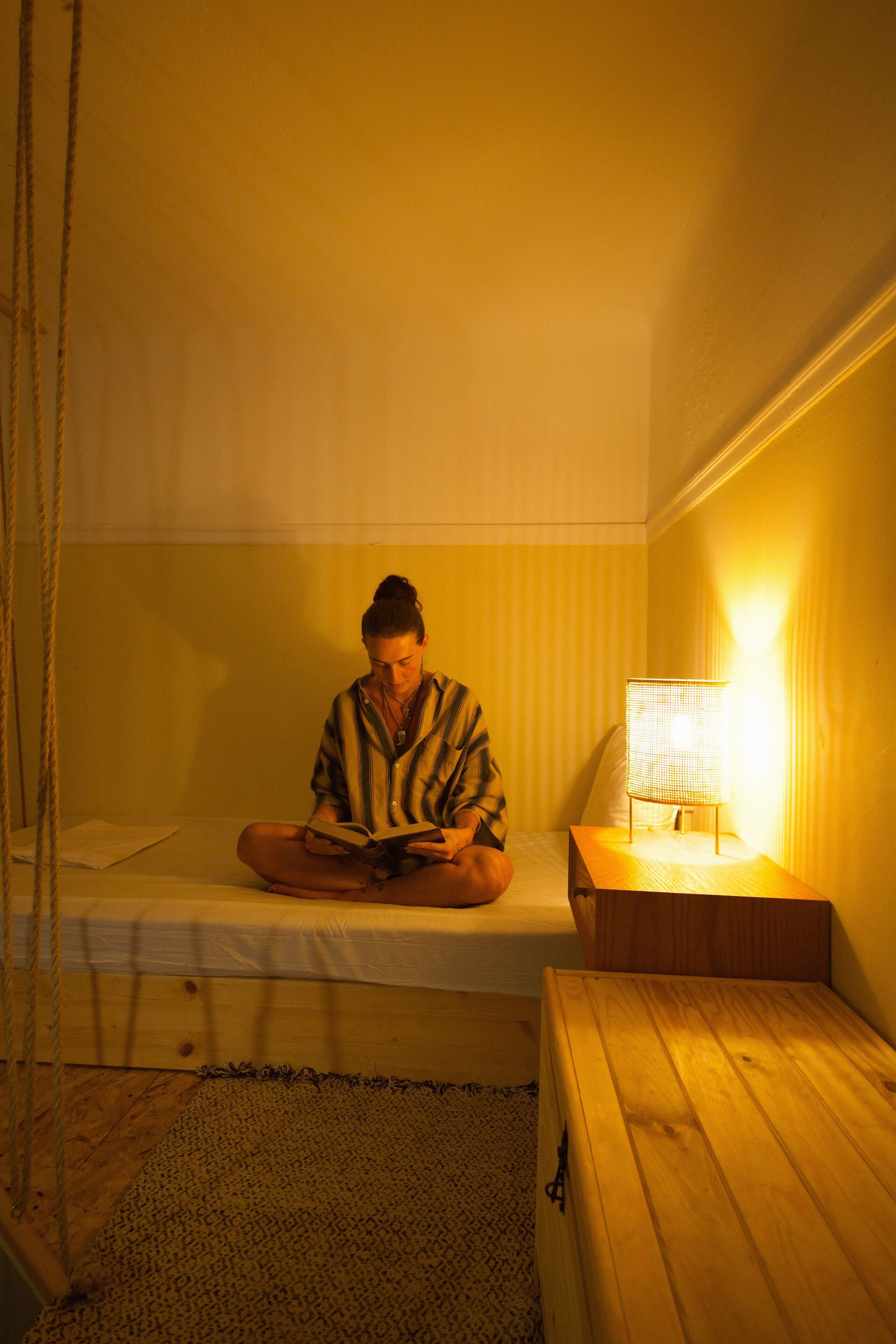 Eine Person sitzt auf einem Bett in einem Zimmer mit einem Nachttisch aus Holz und einer Lampe.