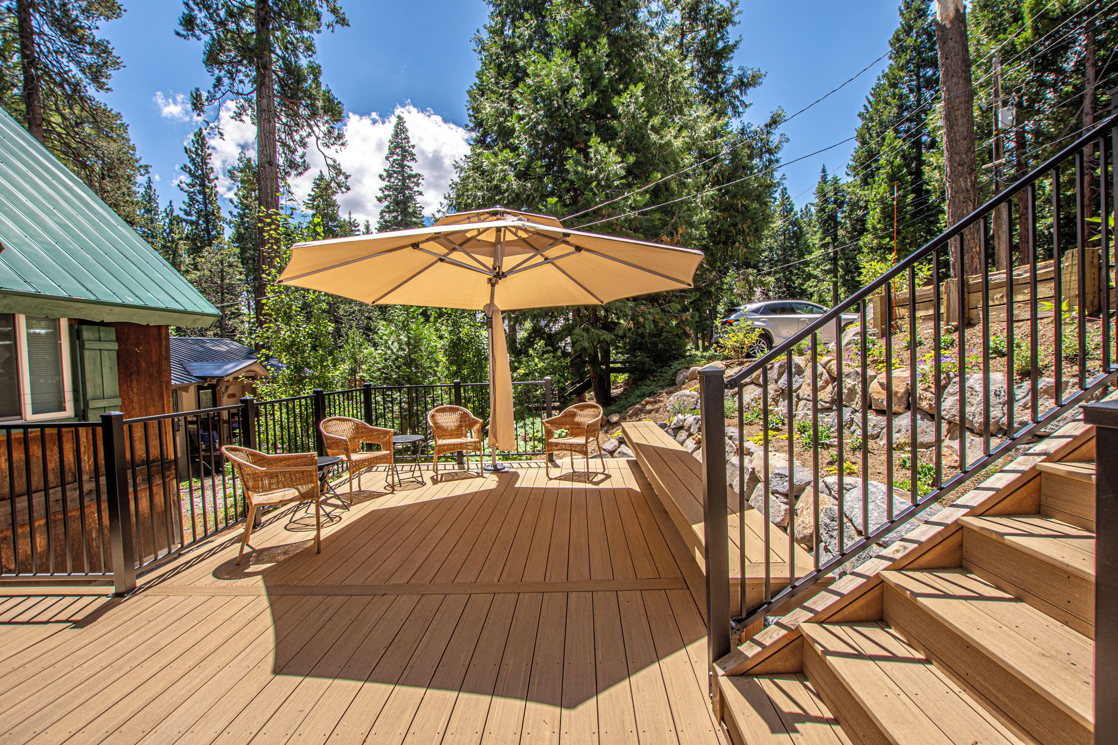 Una terraza de madera con mobiliario exterior, un paraguas grande y una escalera que lleva a un nivel superior.