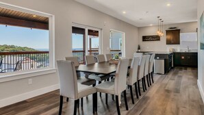 A spacious dining area with a long table and multiple chairs, a kitchen with modern appliances, and large windows offering a view of the outdoors. - Next to Indoor and Outdoor Pools! Hot Tub! Great views! 2 Living Rooms (Branson)