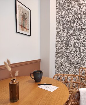 Une table ronde en bois, sur laquelle se trouvent une tasse en céramique foncée, un stylo et une feuille de papier. Un vase contenant des herbes de la pampa séchées est posé sur la table. Un tableau encadré est accroché au mur, au-dessus de la table.