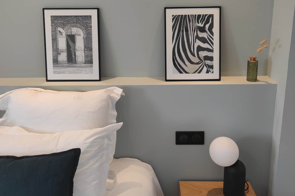 Een slaapkamer met een bed, kussens, een nachtkastje met een lamp en twee ingelijste foto's aan de muur.