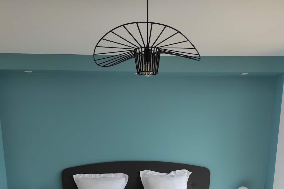 Een net opgemaakt bed met wit en mosterdgekleurde beddengoed, een zwart hoofdeinde en een bijzondere zwarte hanglamp.