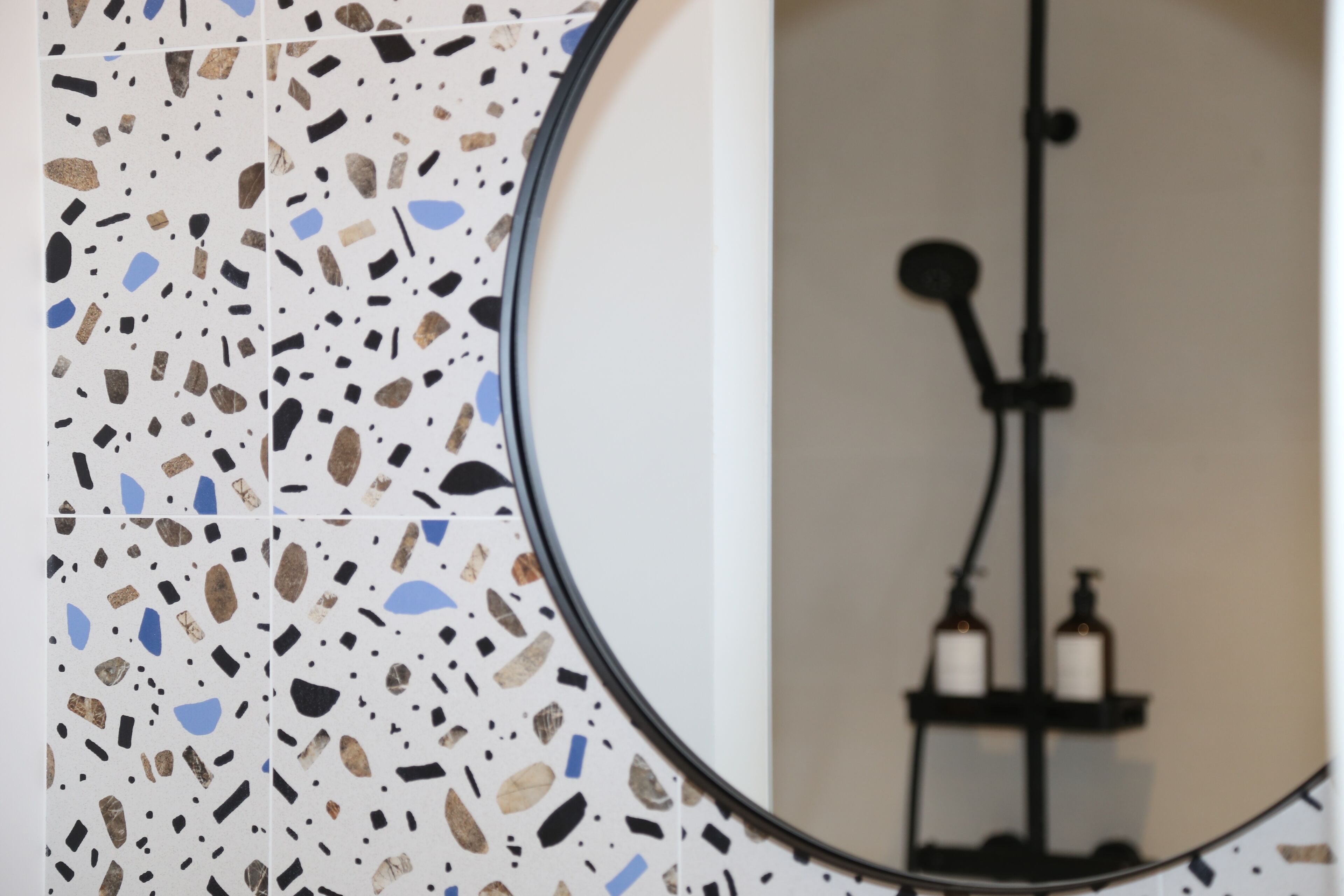 A bathroom with a terrazzo floor, a round mirror, and a towel rack with bottles.