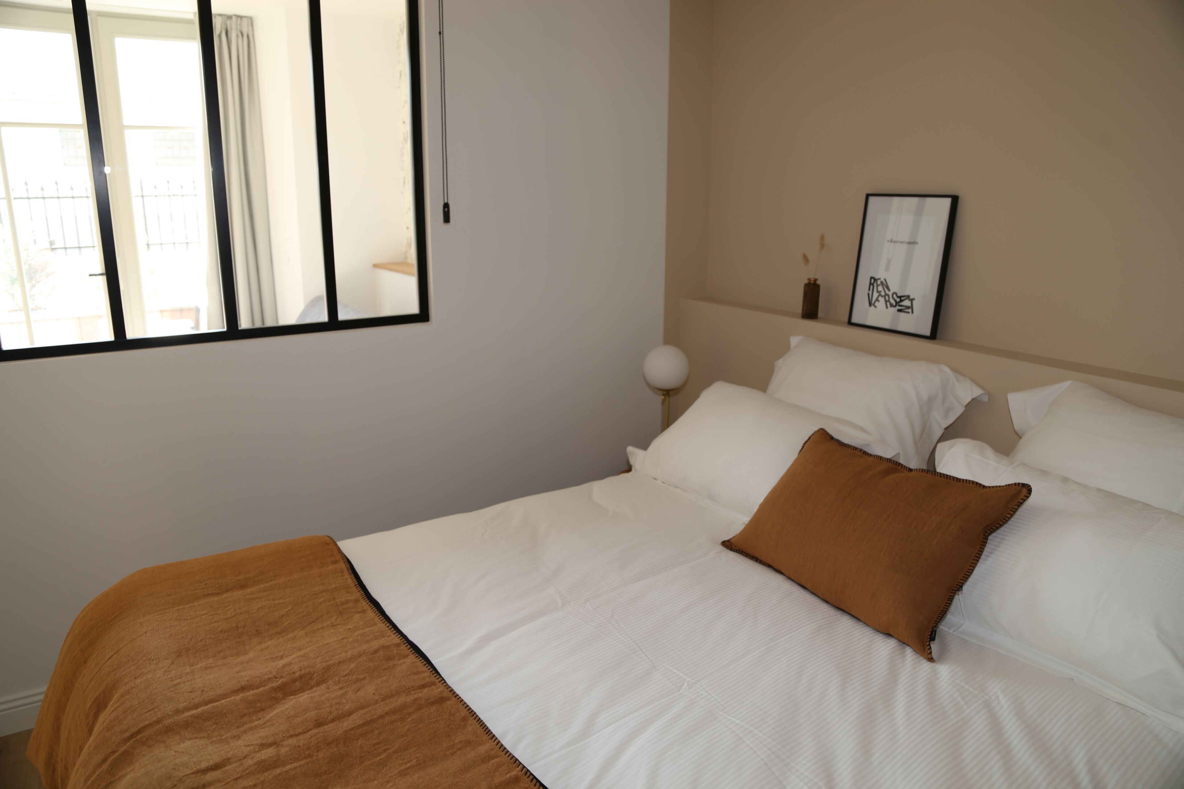 Una cama bien tendida con sábanas blancas y un plaid marrón, una mesita de noche con una lámpara y un cuadro en la pared.