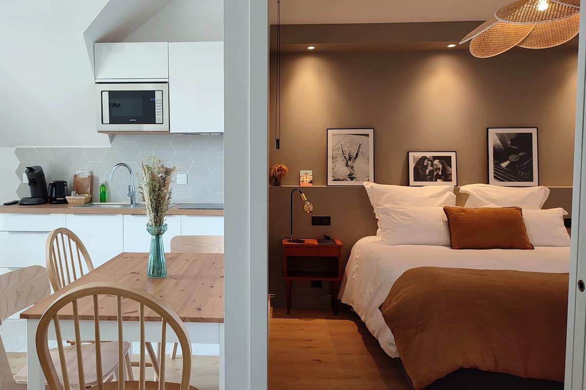 Een moderne keuken met een magnetron, koffiezetapparaat en een vaas met gedroogde bloemen. Grenzend hieraan een slaapkamer met een bed, kussens en ingelijste foto's aan de muur.