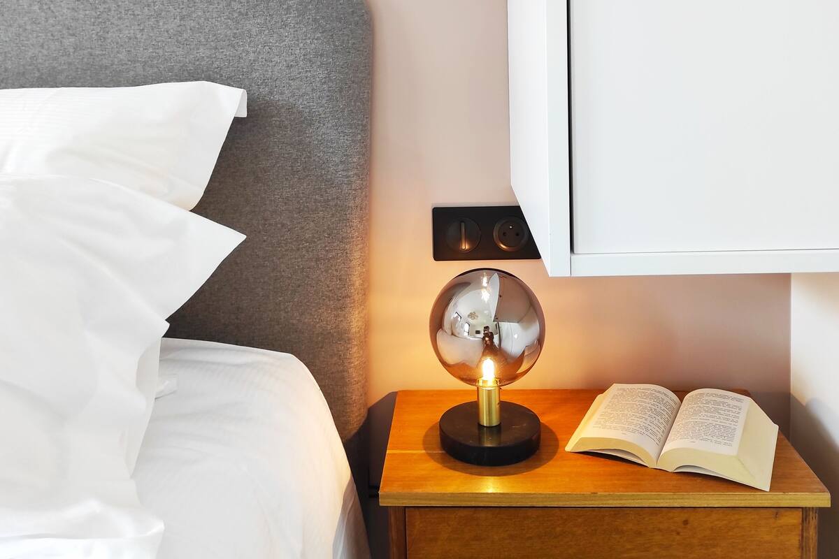 Een keurig opgemaakt bed met witte lakens, een nachtkastje met een boek, een lamp en een garderobe in een slaapkamer.