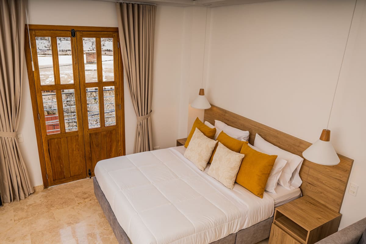 Un dormitorio ordenado con una cama grande, cabecera de madera, dos mesitas de noche con lámparas y una puerta de madera con paneles de vidrio.